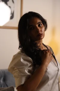 Modelling image of Reshma Das. A lady standing in a dimly warm lighted setting with free hair and in white shirt costume